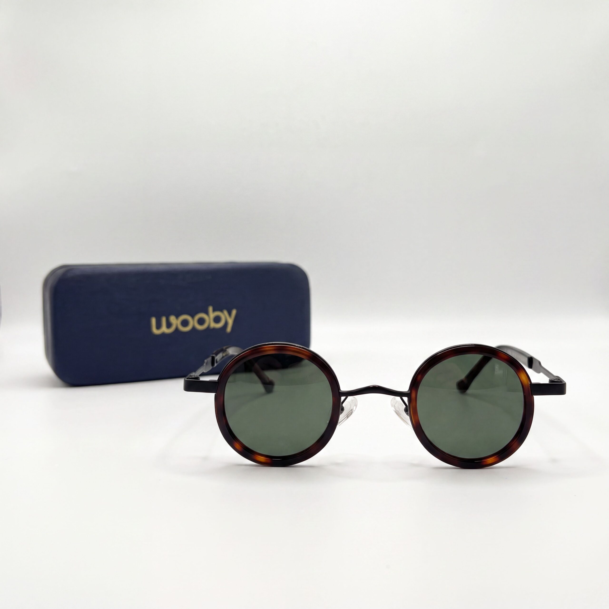 نظارات شمسية دائرية كلاسيكية من ماركة ووبي (Wooby) بإطار بلون الهافان البني والأسود مع عدسات داكنة بجانب علبة زرقاء داكنة